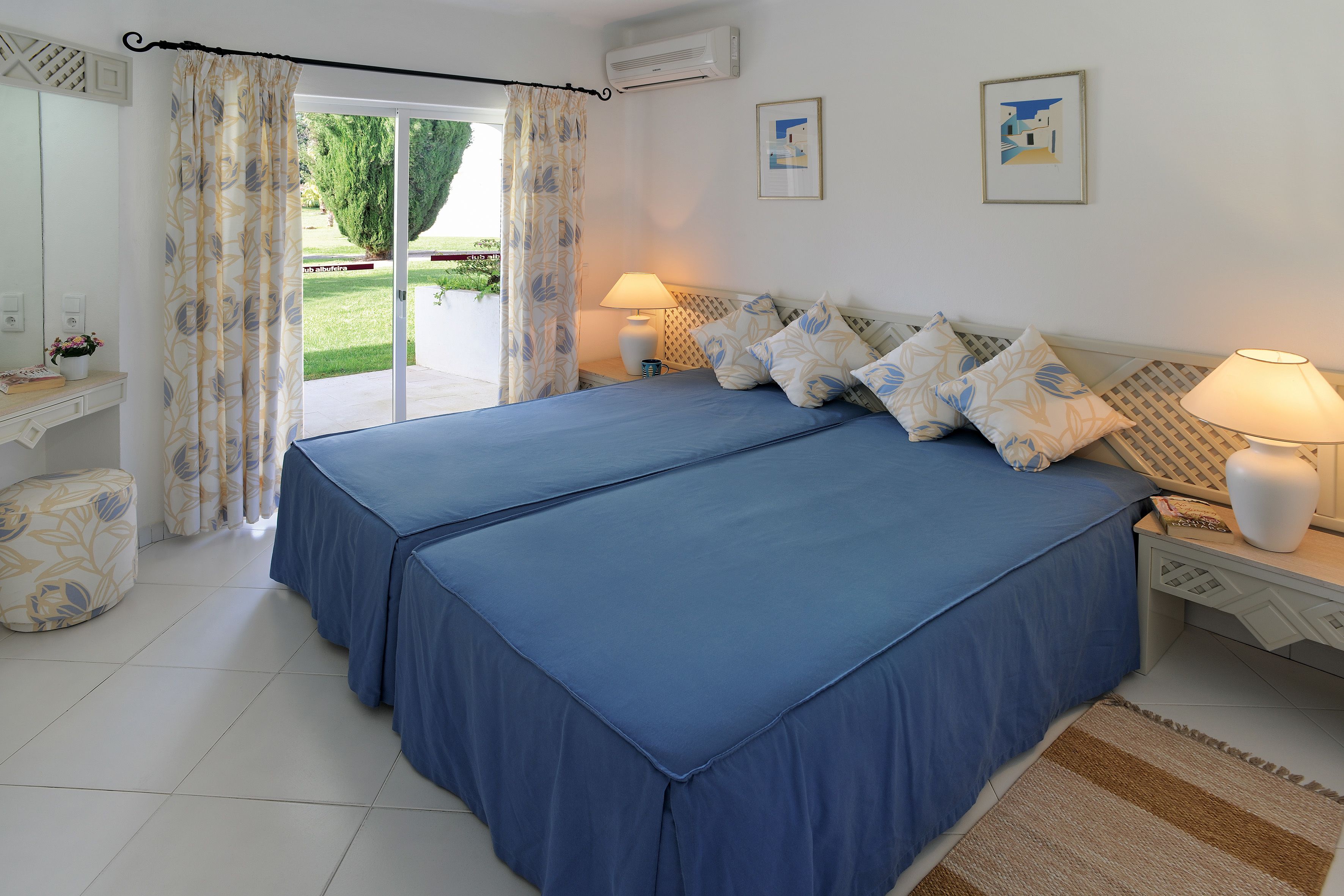 Een slaapkamer met een tweepersoonsbed, twee bedlampjes, een klein tafeltje met een lamp en uitzicht op een tuin door een glazen deur.