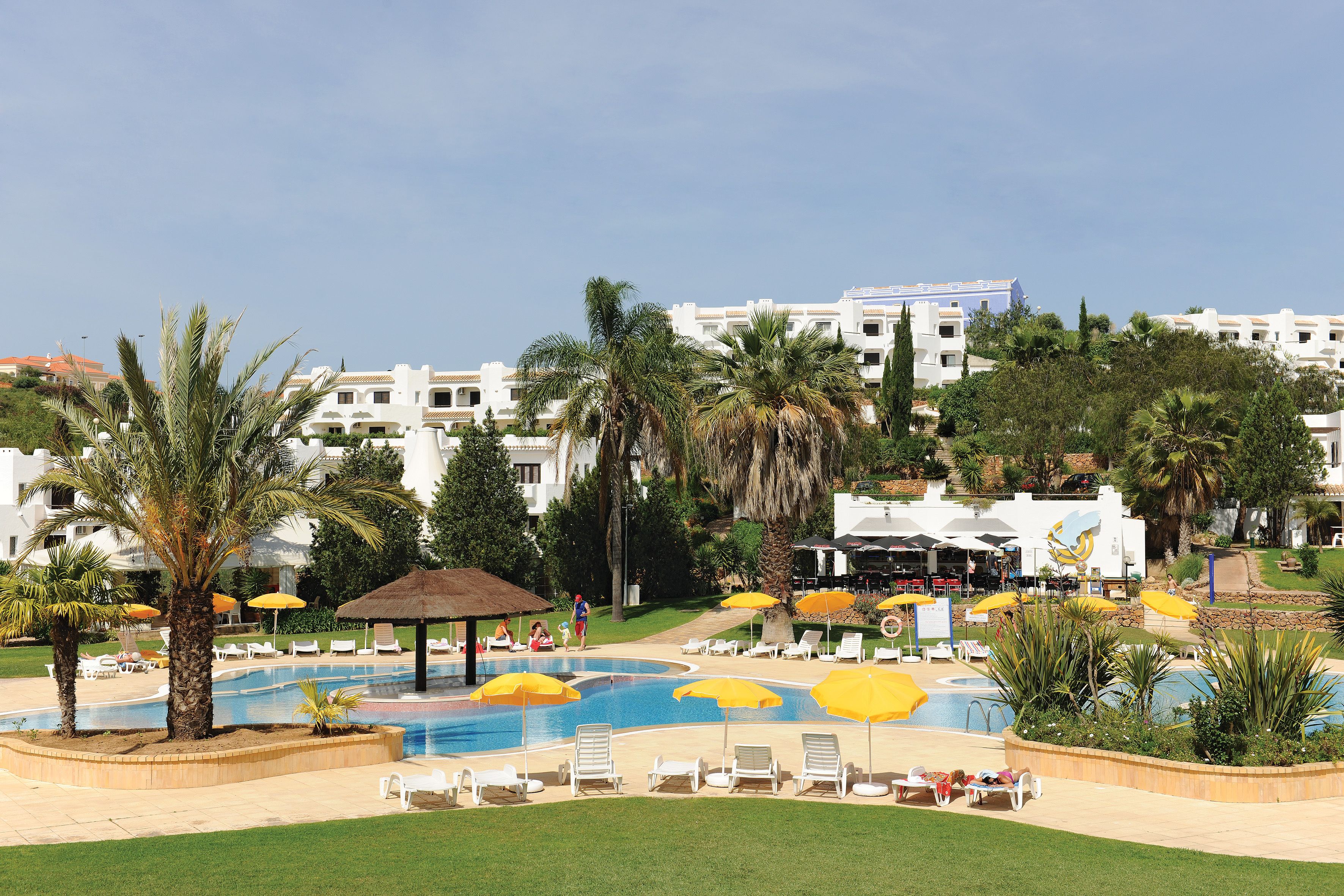 Ein Poolbereich eines Resorts mit mehreren Liegestühlen und Sonnenschirmen, umgeben von Palmen und einem Gebäude im Hintergrund.