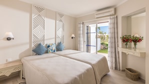 Una cama doble con colcha color beige, una mesita de noche con lámpara, un panel decorativo en la pared y vistas al exterior a través de una puerta de cristal.