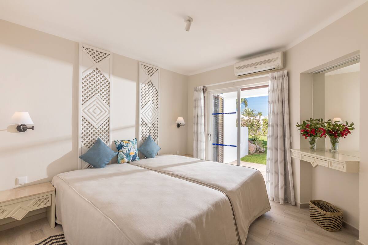 Ein Doppelbett mit beige farbenem Bettbezug, ein Nachttisch mit Lampe, eine dekorative Wandverkleidung und Blick ins Freie durch eine Glastür.