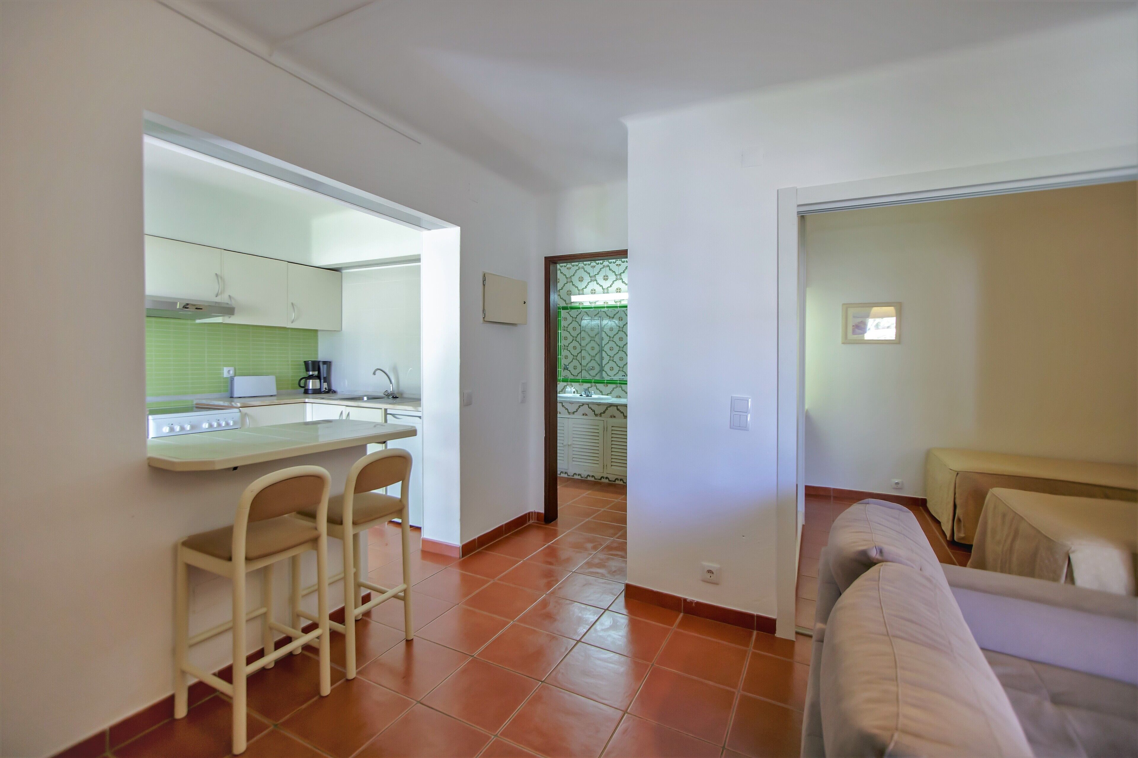 A compact kitchen with a counter and two bar stools, a dining area with a sofa, and a view of a balcony with a green railing.
