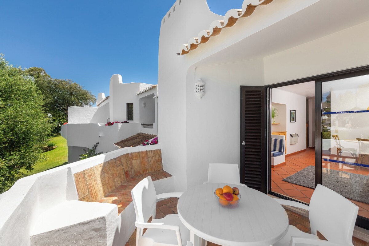 A white outdoor patio with a round table and chairs, a bowl of fruit, and a view of the garden and neighboring buildings.