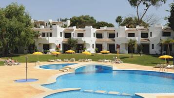 A swimming pool with sun loungers and umbrellas, surrounded by a white building with balconies.