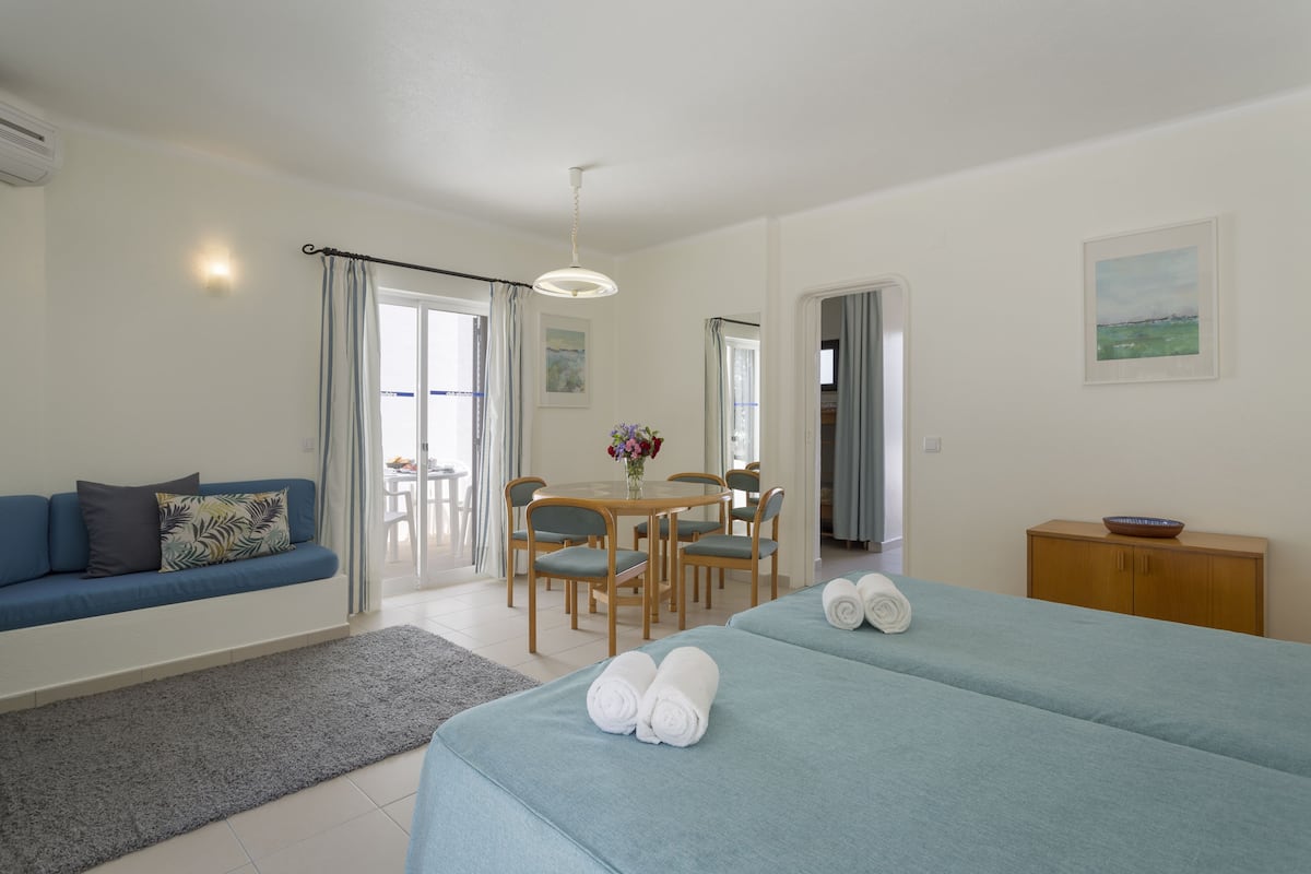Ein Hotelzimmer mit Bett, einer Couch, einem Esstisch mit Stühlen, einem kleinen Schrank und einem Fenster mit Vorhängen.