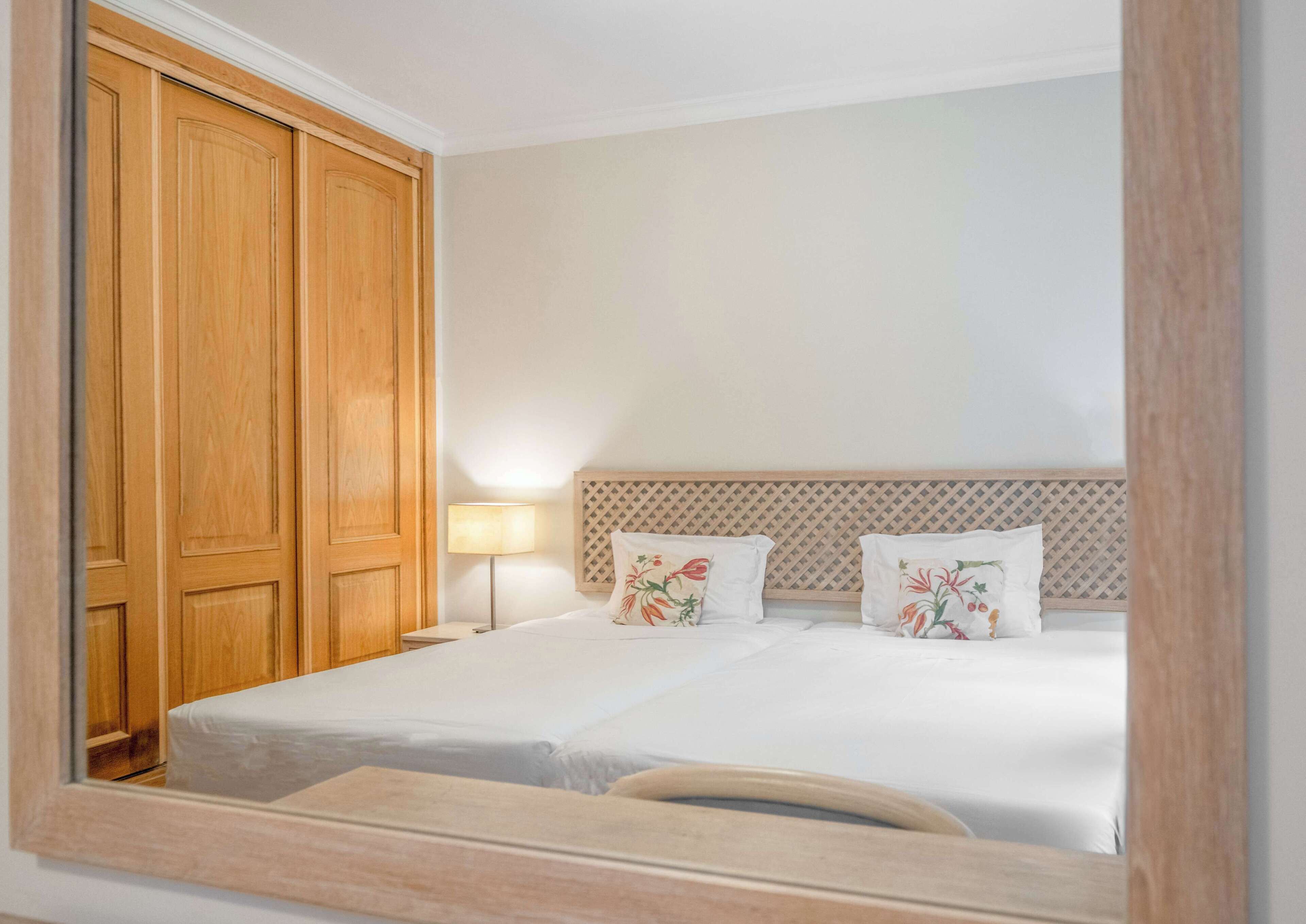 Quarto de hotel com um grande guarda-roupa de madeira, uma cama com almofadas florais e uma mesa de cabeceira com abajur.