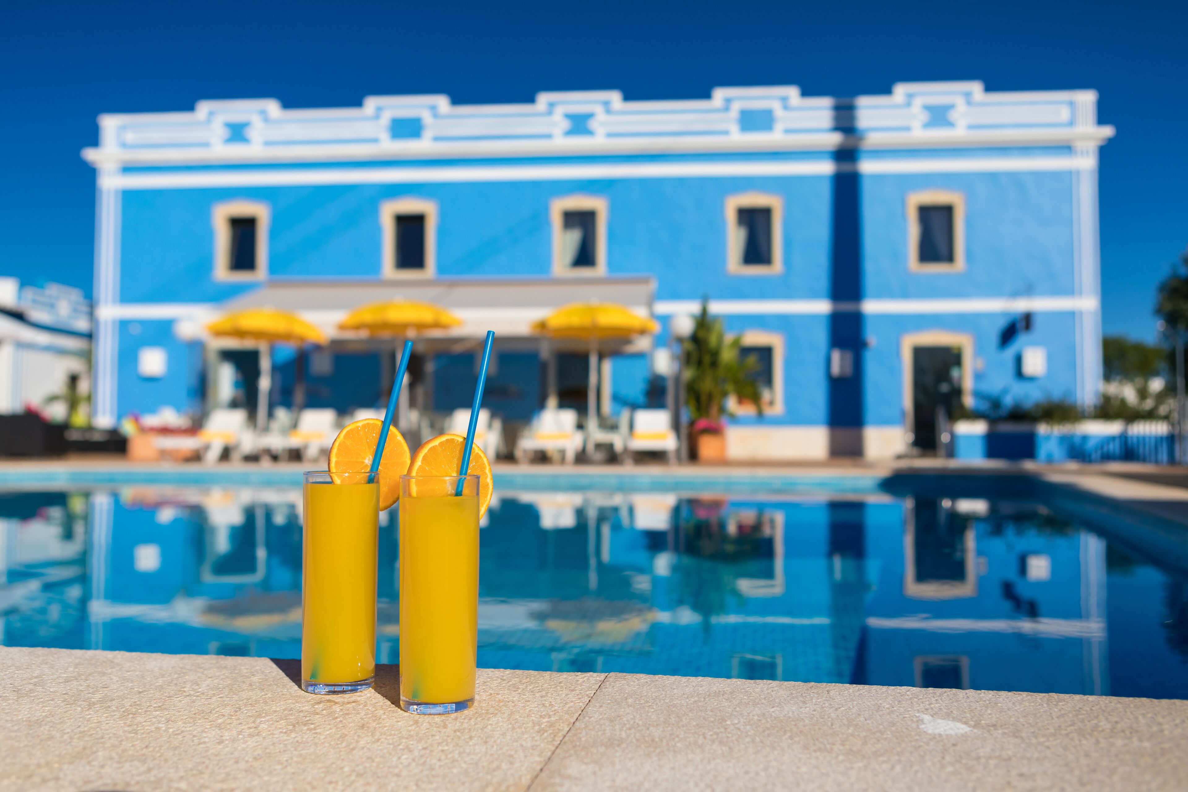 Two tall glasses of orange juice with straws and orange slices on a poolside table, with a blue building and umbrellas in the background.