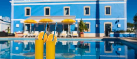 Two tall glasses of orange juice with straws and orange slices on a poolside table, with a blue building and umbrellas in the background.
