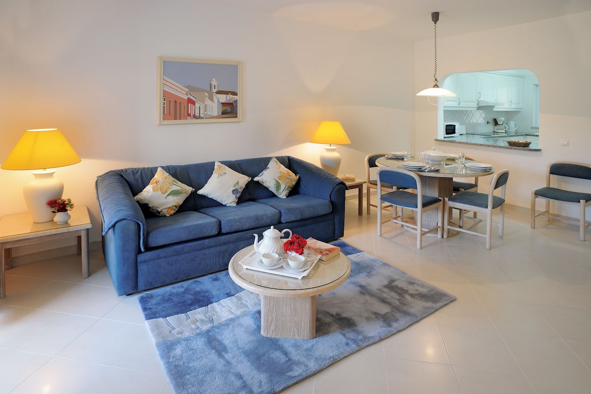 Ein Wohnzimmer mit einem blauen Sofa, einem Couchtisch mit Teekanne sowie einem Essbereich mit Stühlen und Tisch.