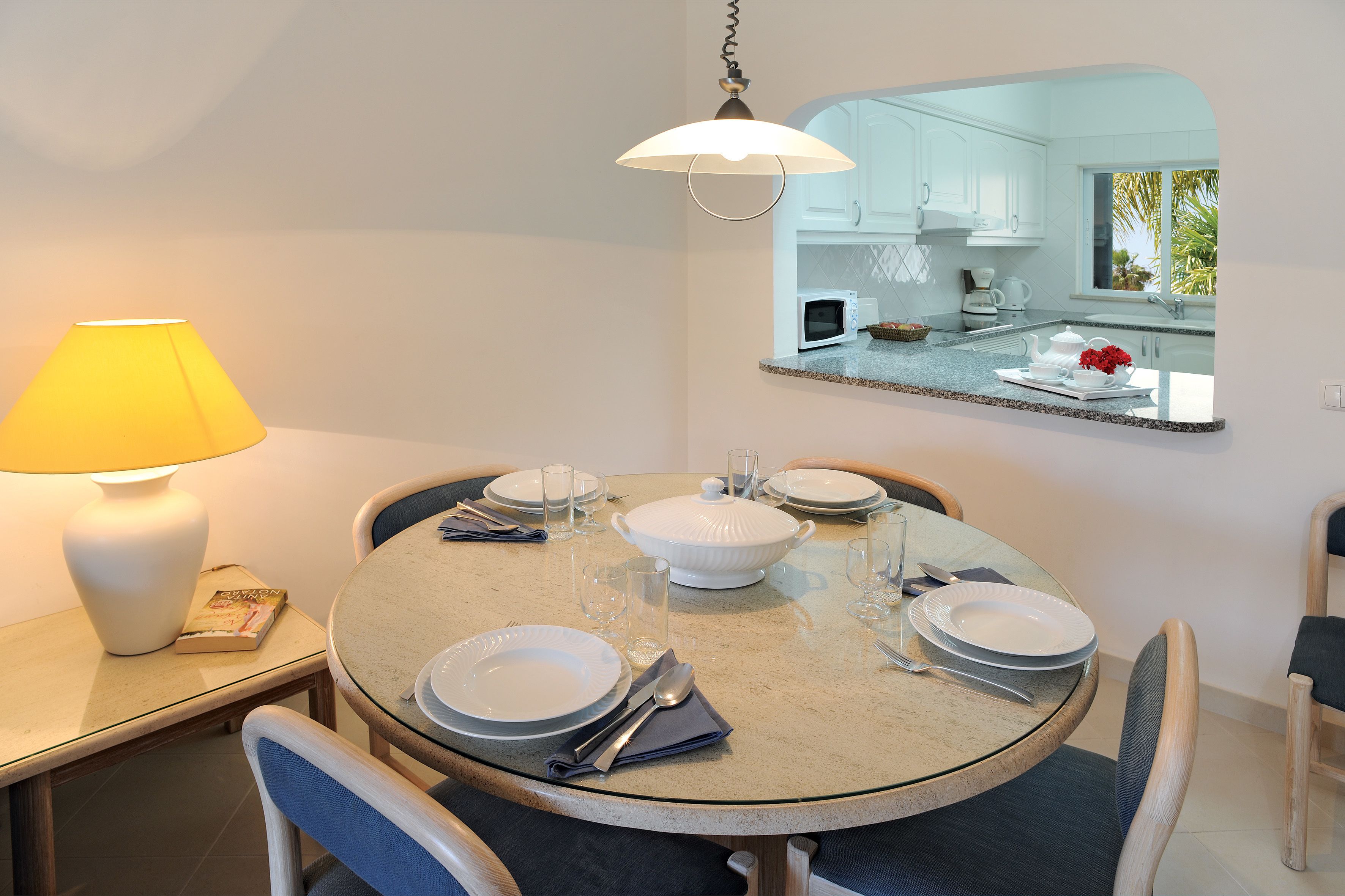 A dining area with a round table set for four, a lamp, and a view of the kitchen through a mirror.