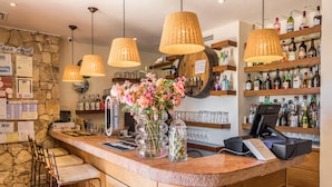 Una zona de bar con pared de piedra, mostrador de madera, taburetes y una variedad de botellas y vasos en estantes.