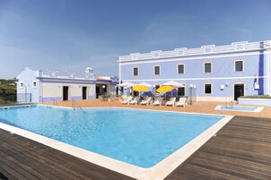 Una piscina con sillas de descanso y sombrillas, una terraza de madera y un edificio con fachada azul.