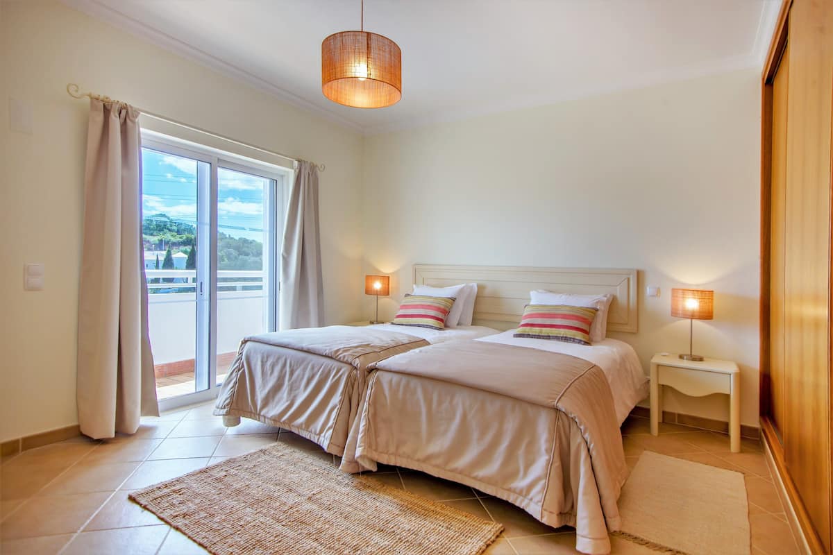 Ein Schlafzimmer mit zwei Betten, einem großen Fenster mit Vorhängen, einer Deckenleuchte und einem Nachttisch mit Lampe.