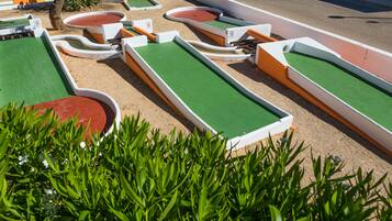 Een minigolfbaan met meerdere groene en rode gaten, omgeven door zand en begrensd door witte en oranje constructies.
