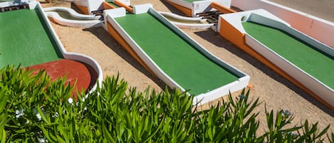 A miniature golf course with multiple green and red holes, surrounded by sand and bordered by white and orange structures.