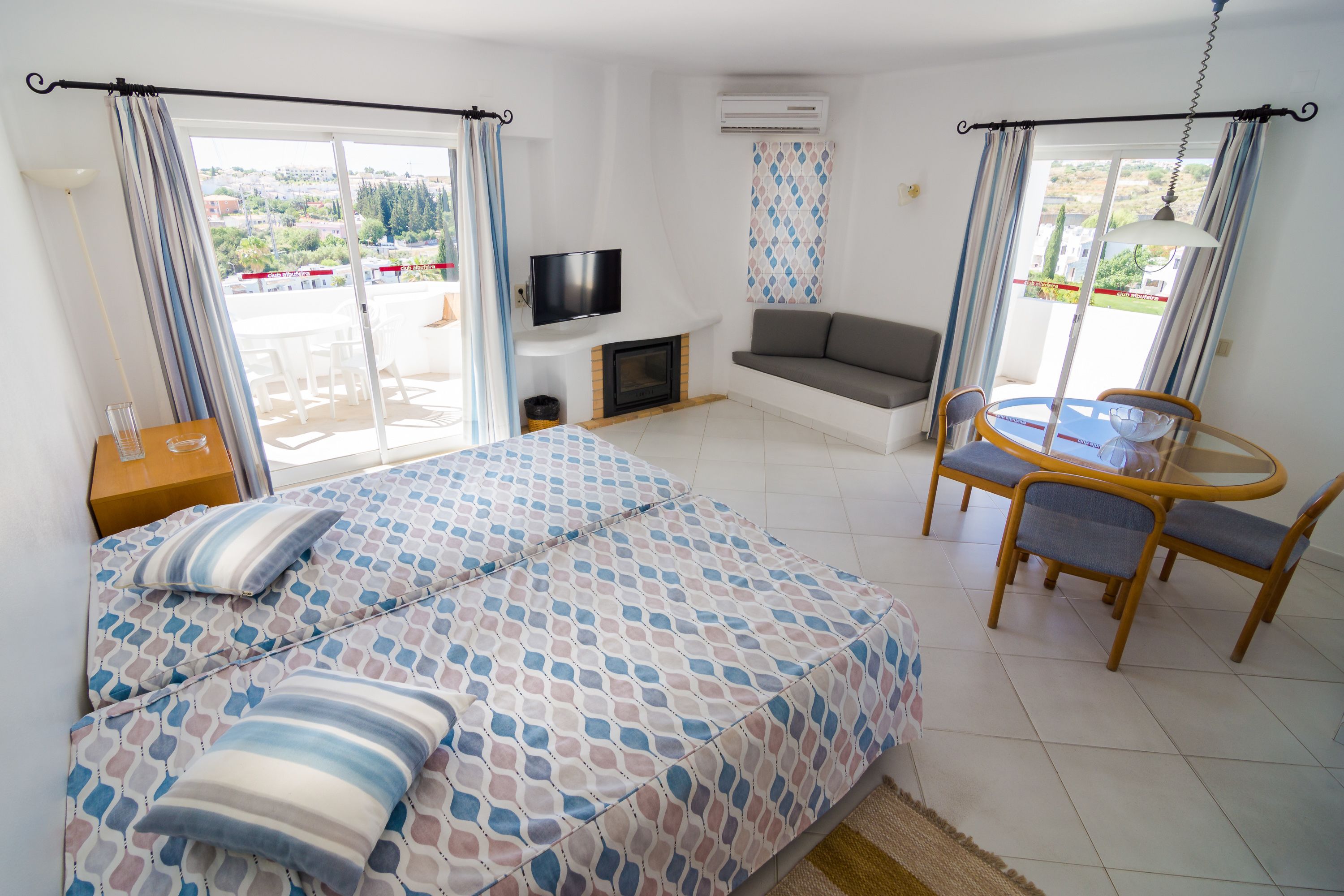 Ein Schlafzimmer mit Bett, Fernseher, Sofa, Esstisch und Stühlen sowie Blick ins Freie.