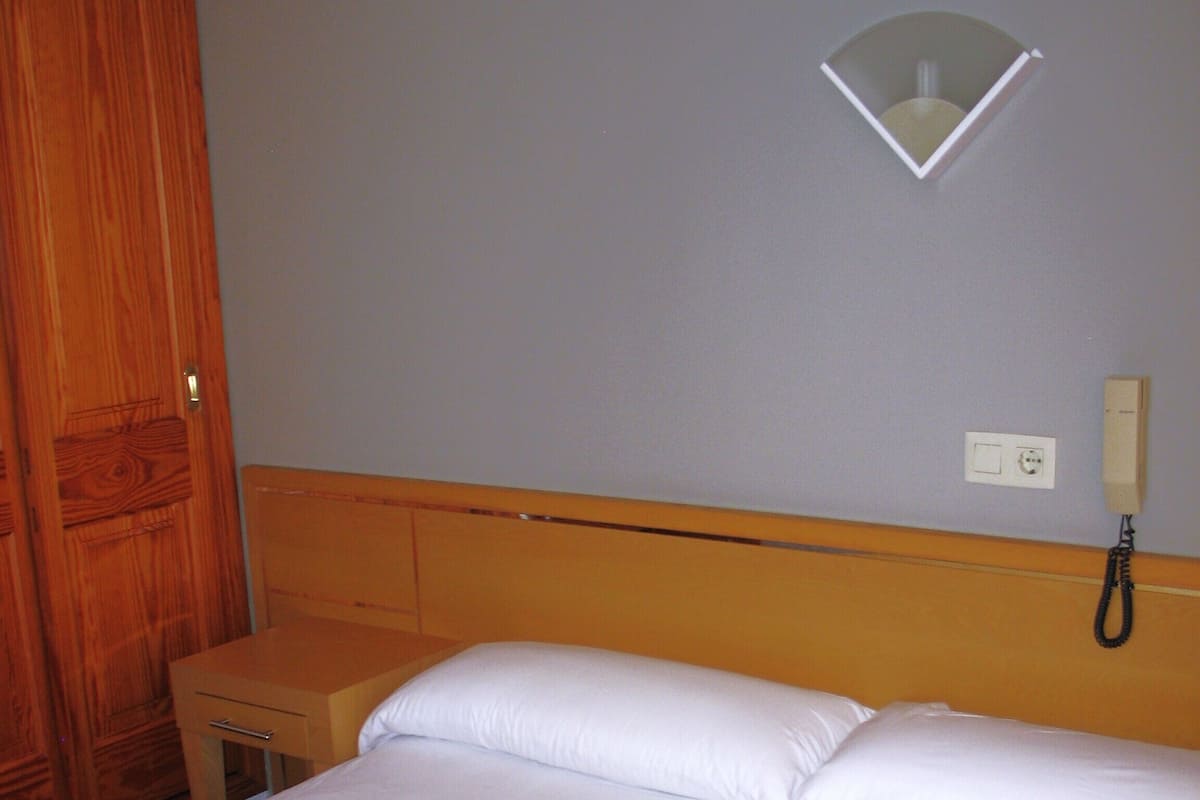 A neatly made bed with white linens, a wooden headboard, a bedside table, and a wall-mounted telephone.