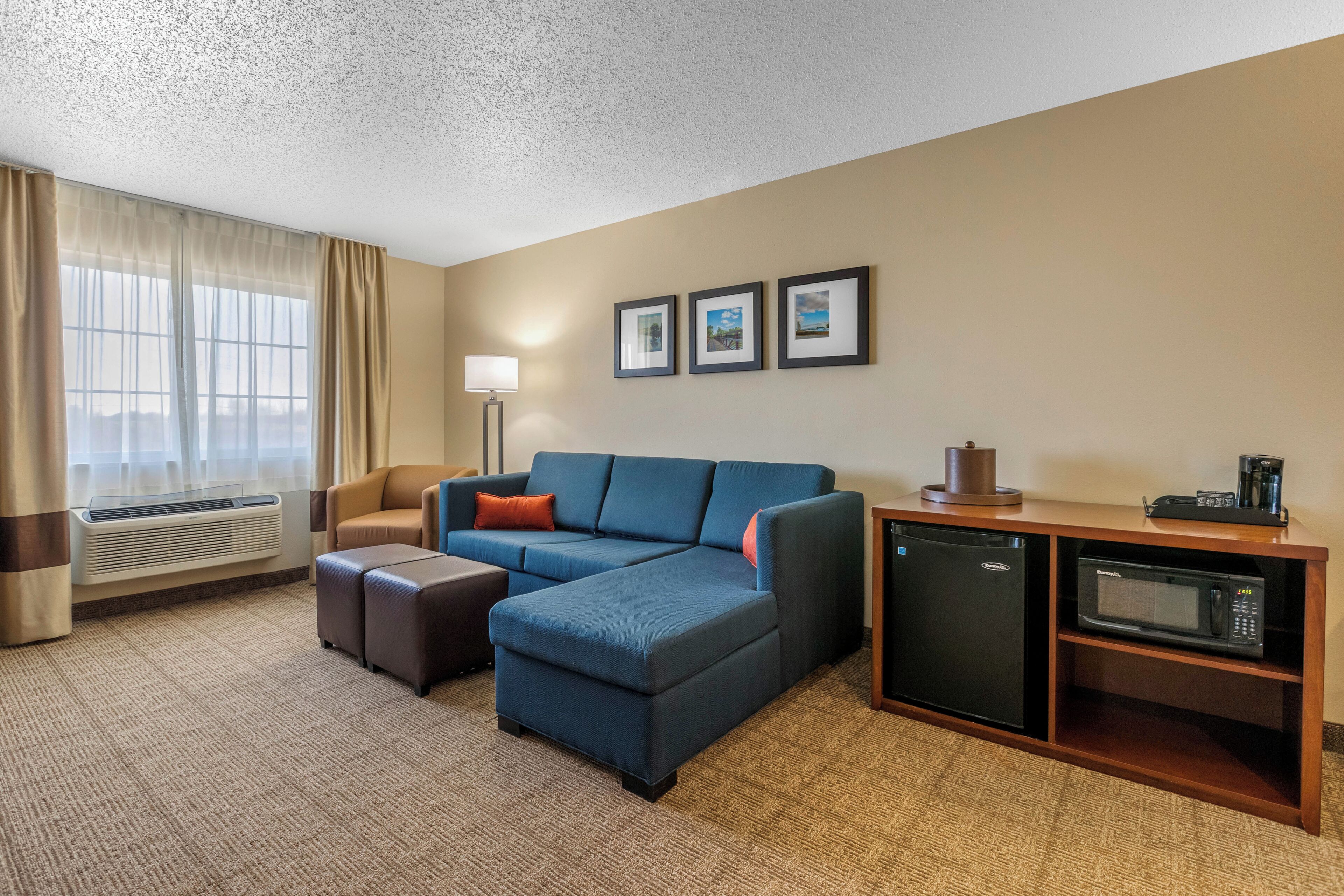 A hotel room with a sectional sofa, a small kitchenette with a microwave, a refrigerator, and a window with curtains.