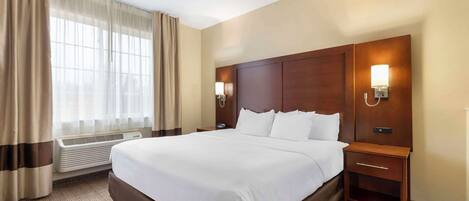 A hotel room with a large bed, a wooden headboard, two bedside tables with lamps, a window with curtains, and a wall-mounted air conditioning unit.
