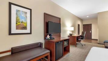A hotel room with a bed, a desk with a television, a refrigerator, a microwave, a chair, and a bench.