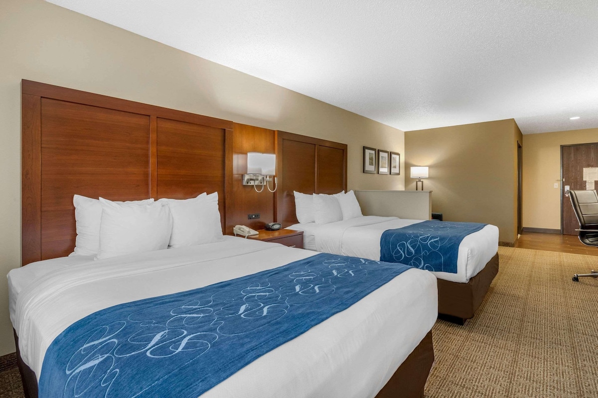 A hotel room with two beds, a wooden headboard, a bedside table with a lamp, and a chair.