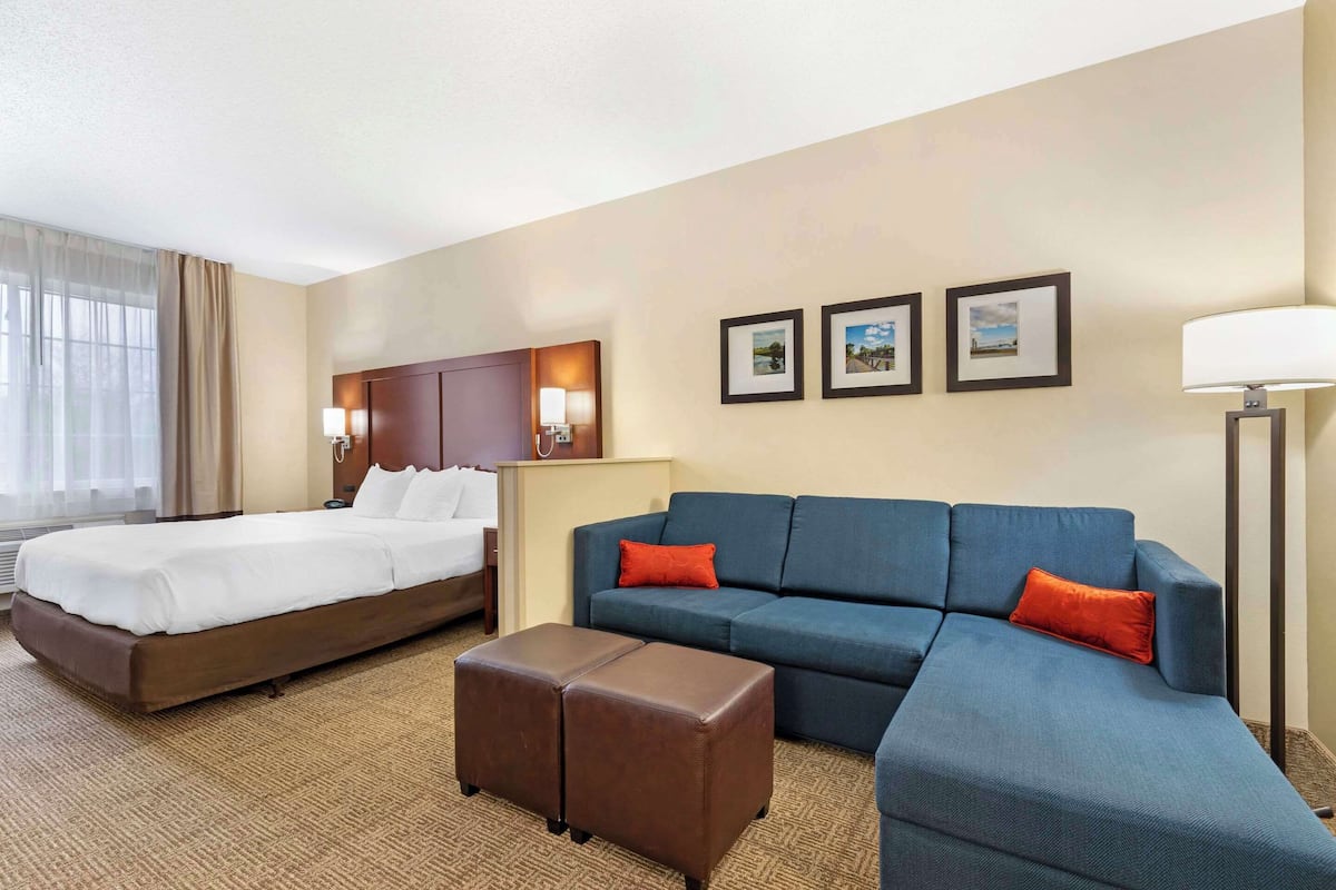 Una camera d'albergo con un letto, una poltrona, un poggiapiedi e un comodino con una lampada.