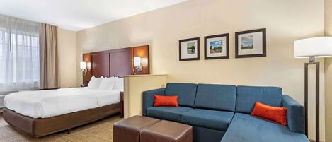 A hotel room with a bed, a sofa, an ottoman, and a nightstand with a lamp.