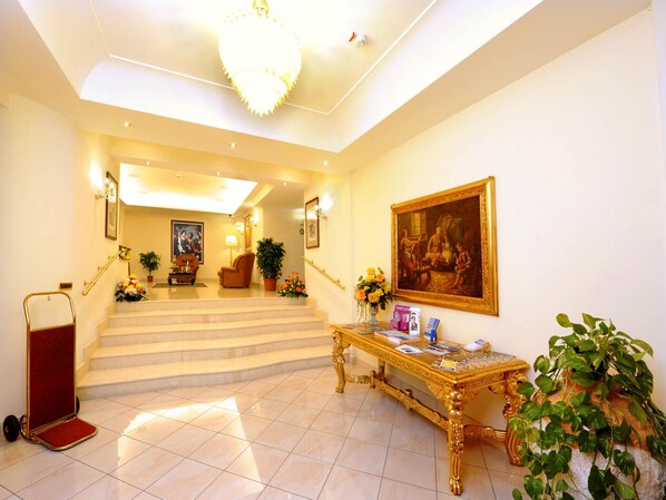 A grand lobby with marble stairs, a chandelier, a painting, and a reception desk. - Buono Hotel (Naples)