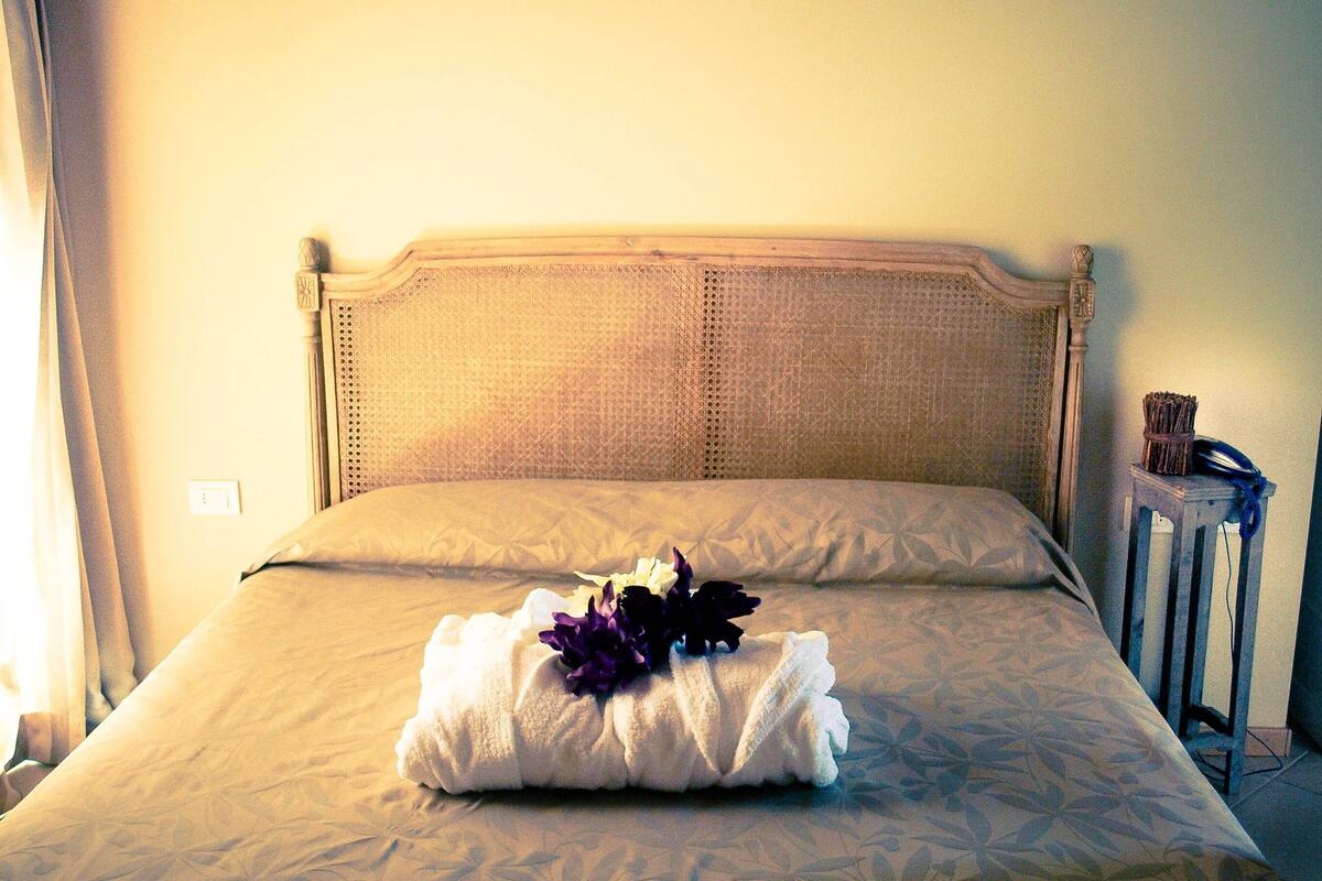A neatly made bed with a floral-patterned bedspread, a folded towel with flowers, and a bedside table with a lamp.