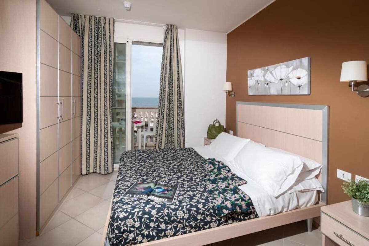 A hotel room with a bed, a TV, a bedside table, a lamp, and a view of the sea through a glass door.