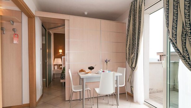 Habitación de hotel moderna con comedor que incluye una mesa y sillas blancas, una pequeña cocina y un balcón con cortinas estampadas.