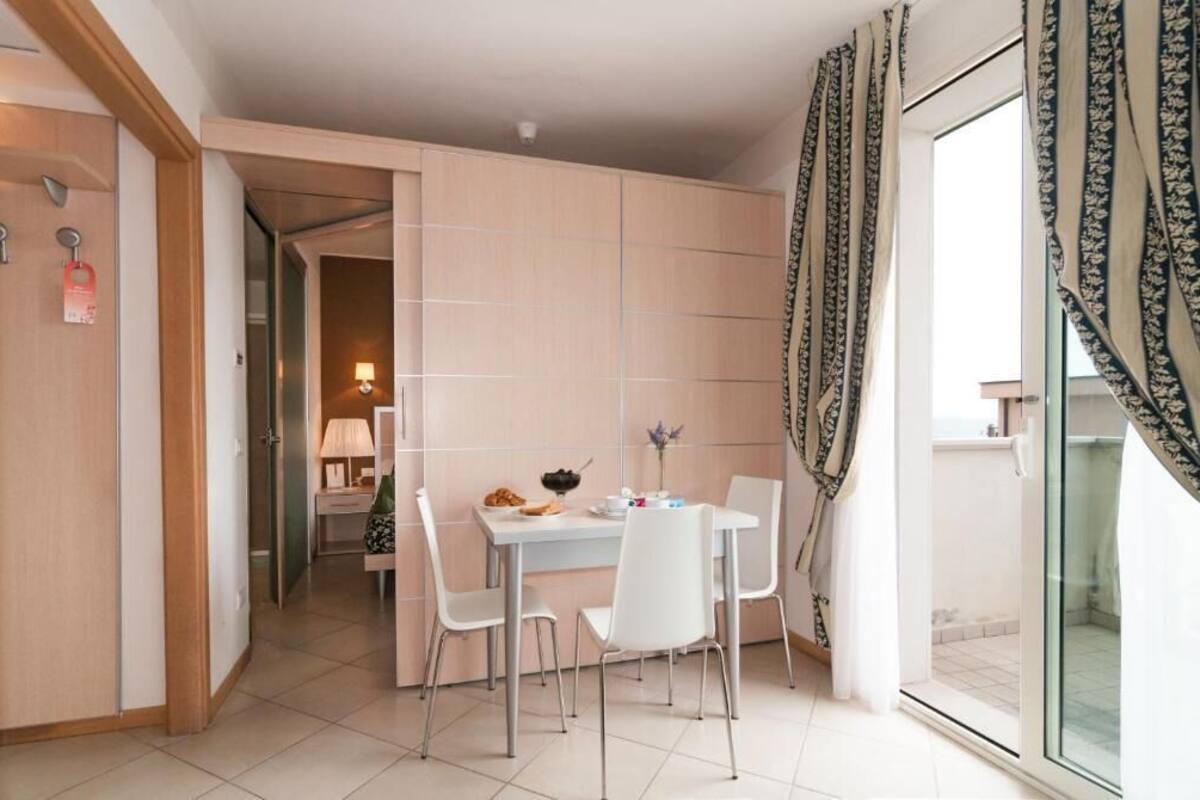 A modern hotel room with a dining area featuring a white table and chairs, a small kitchen area, and a balcony with patterned curtains.