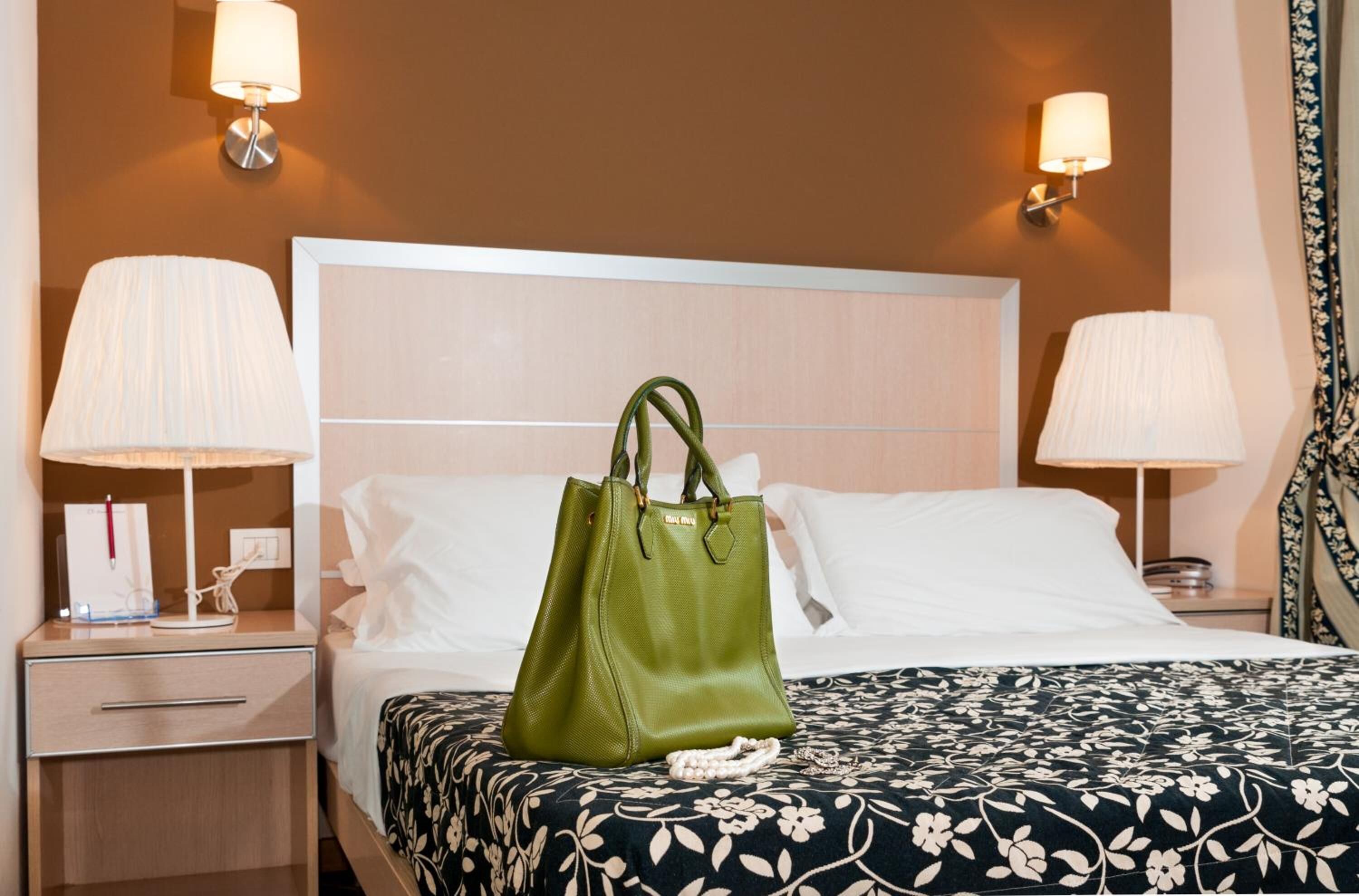 A hotel room with a bed, two bedside lamps, a green handbag on the bed, and a floral bedspread.