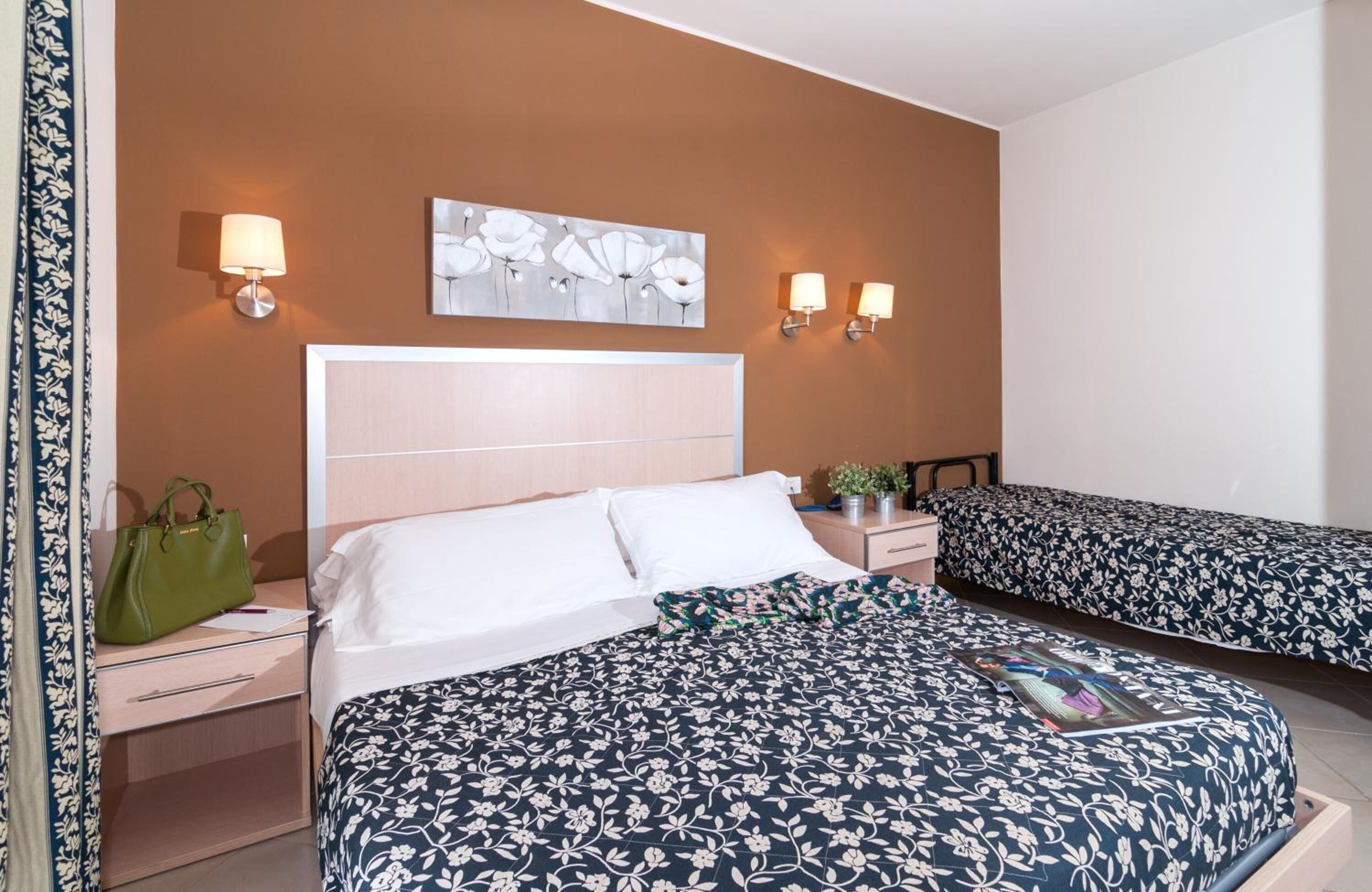 Hotelkamer met een groot bed, nachtkastje met een lamp, een groene handtas, een schilderij aan de muur en een bloemenprint beddeken.