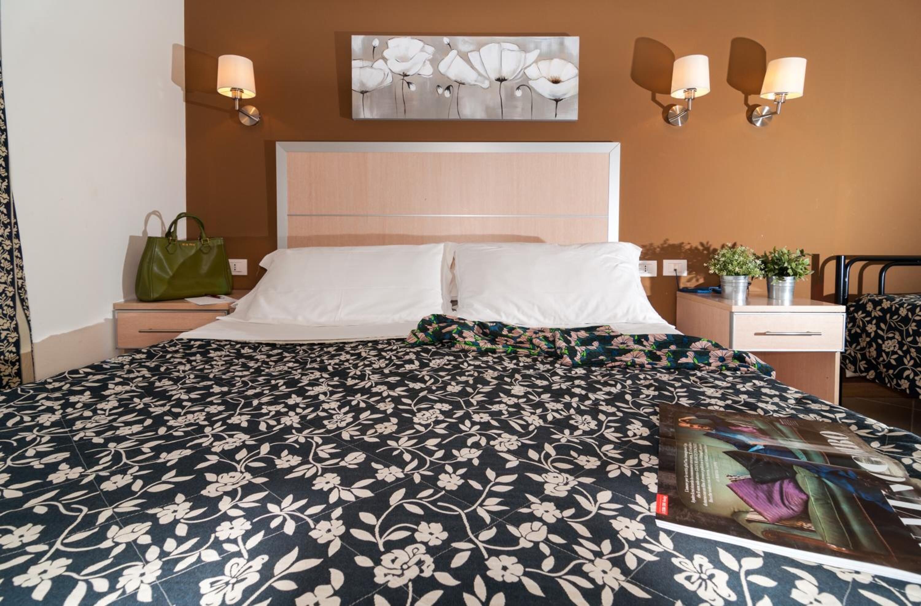 Habitación de hotel con una cama grande, dos mesitas de noche, un bolso verde sobre una de ellas, una lámpara de pared y un cuadro encima de la cama.