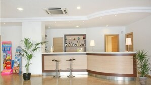 Uma área de recepção com balcão, dois banquetes, uma prateleira com garrafas e plantas em vasos.