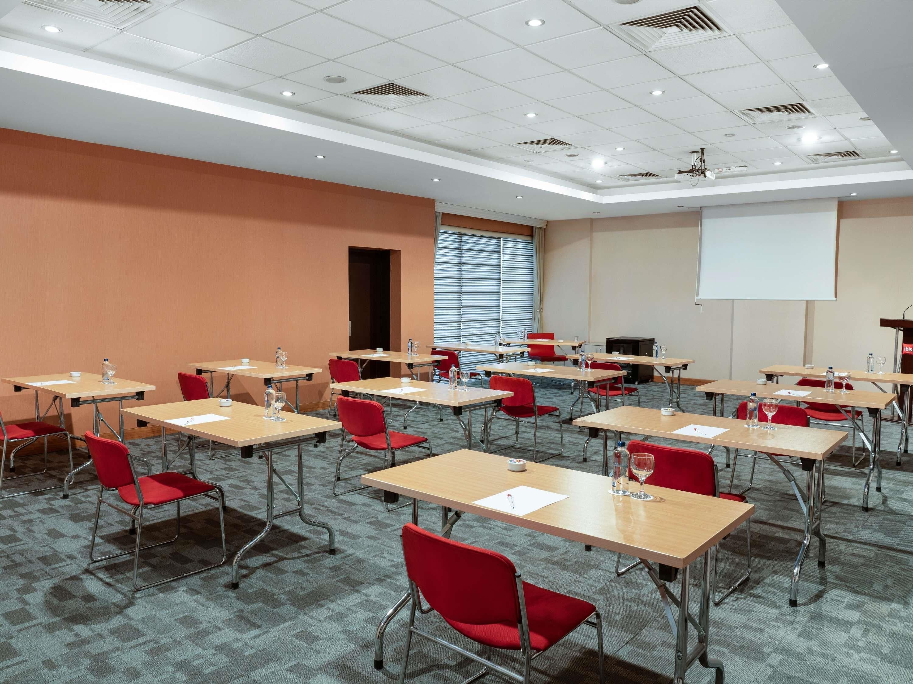 A conference room with multiple tables and chairs, a projector screen, and a large window with blinds.
