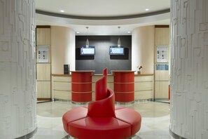 A modern reception area with a central red seating area, two digital information screens on the wall, and a wooden counter.