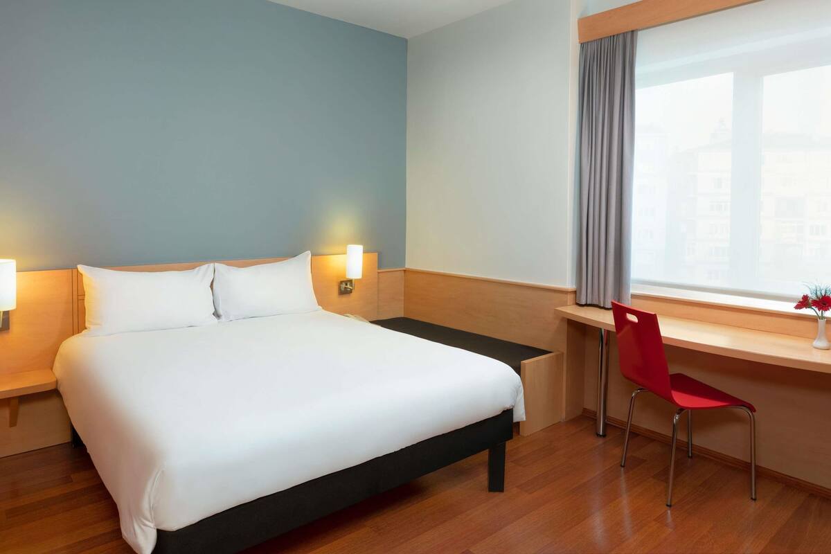 Quarto de hotel com cama, abajures, escrivaninha com cadeira e janela com cortinas.