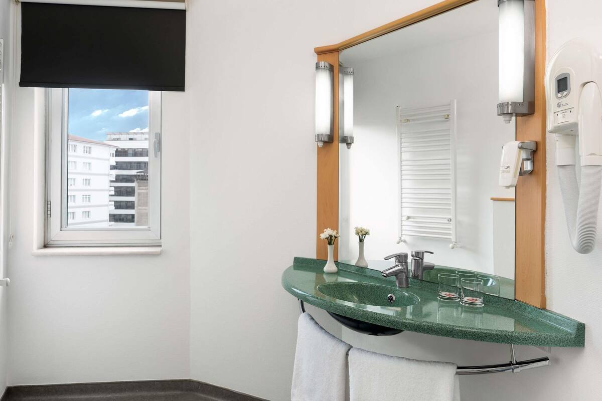Une salle de bain moderne avec un lavabo en verre vert, un grand miroir, un porte-serviettes, une fenêtre avec un store noir et un mur blanc.