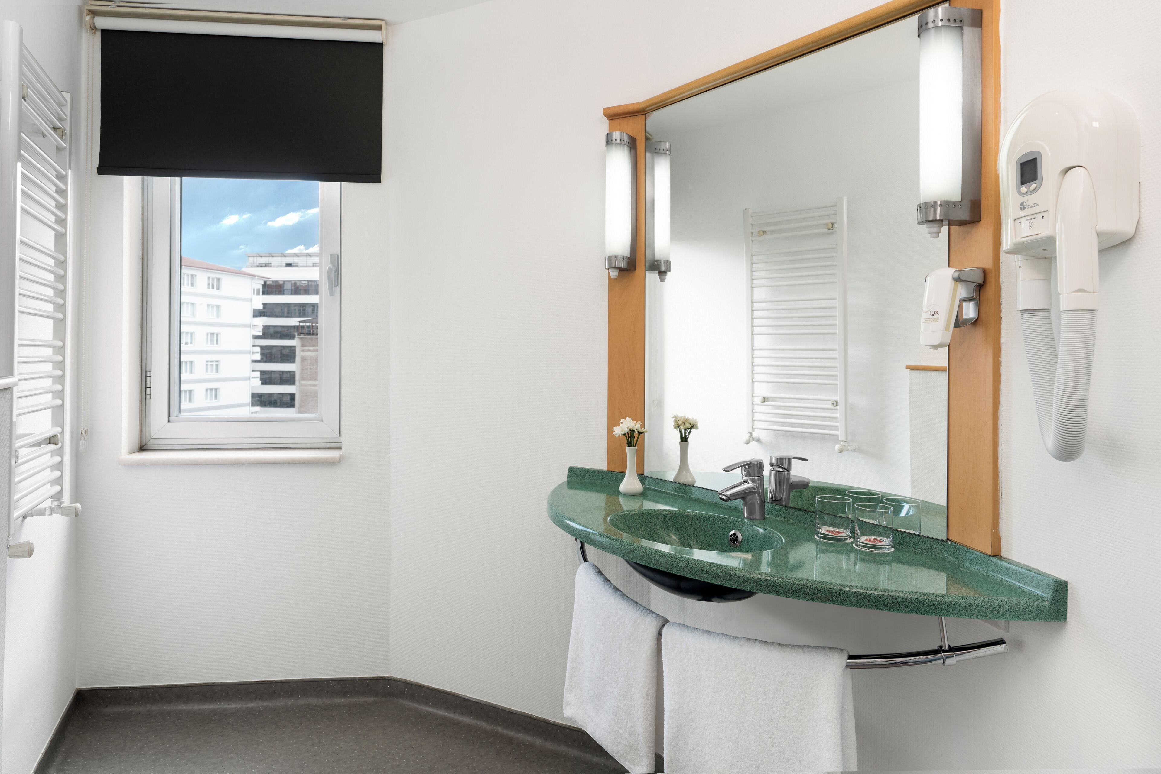 A bathroom with a green sink, a mirror, a towel rack, a window with blinds, and a hairdryer on the wall.