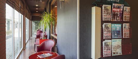 Un pasillo con sillas y mesas rojas, un mueble con revistas en la pared y ventanas amplias a un lado.