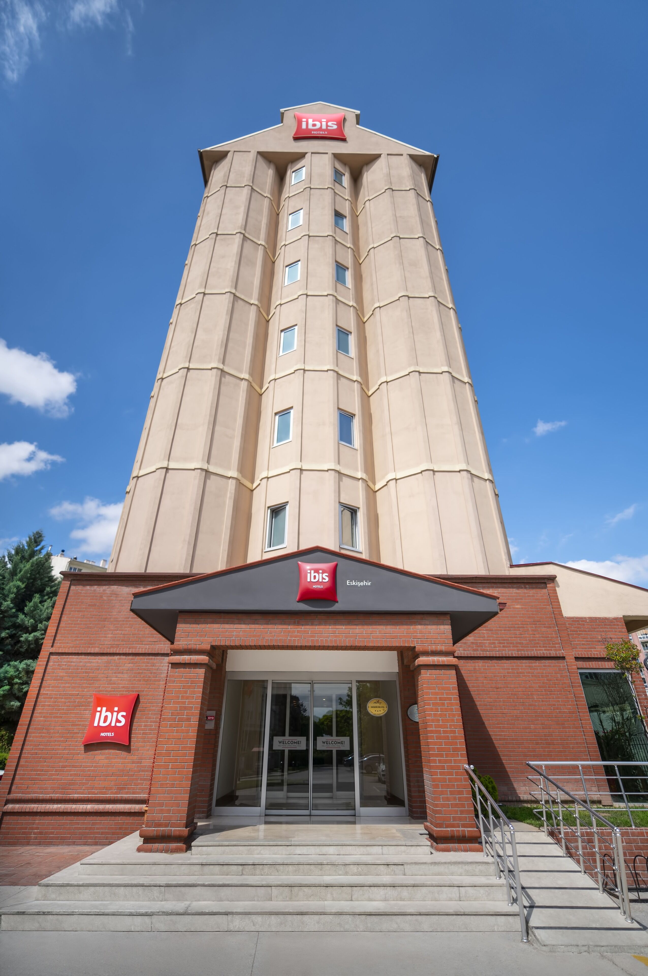 A tall, beige hotel with a red 'ibis' sign on the facade and a 'Welcome' sign at the entrance.