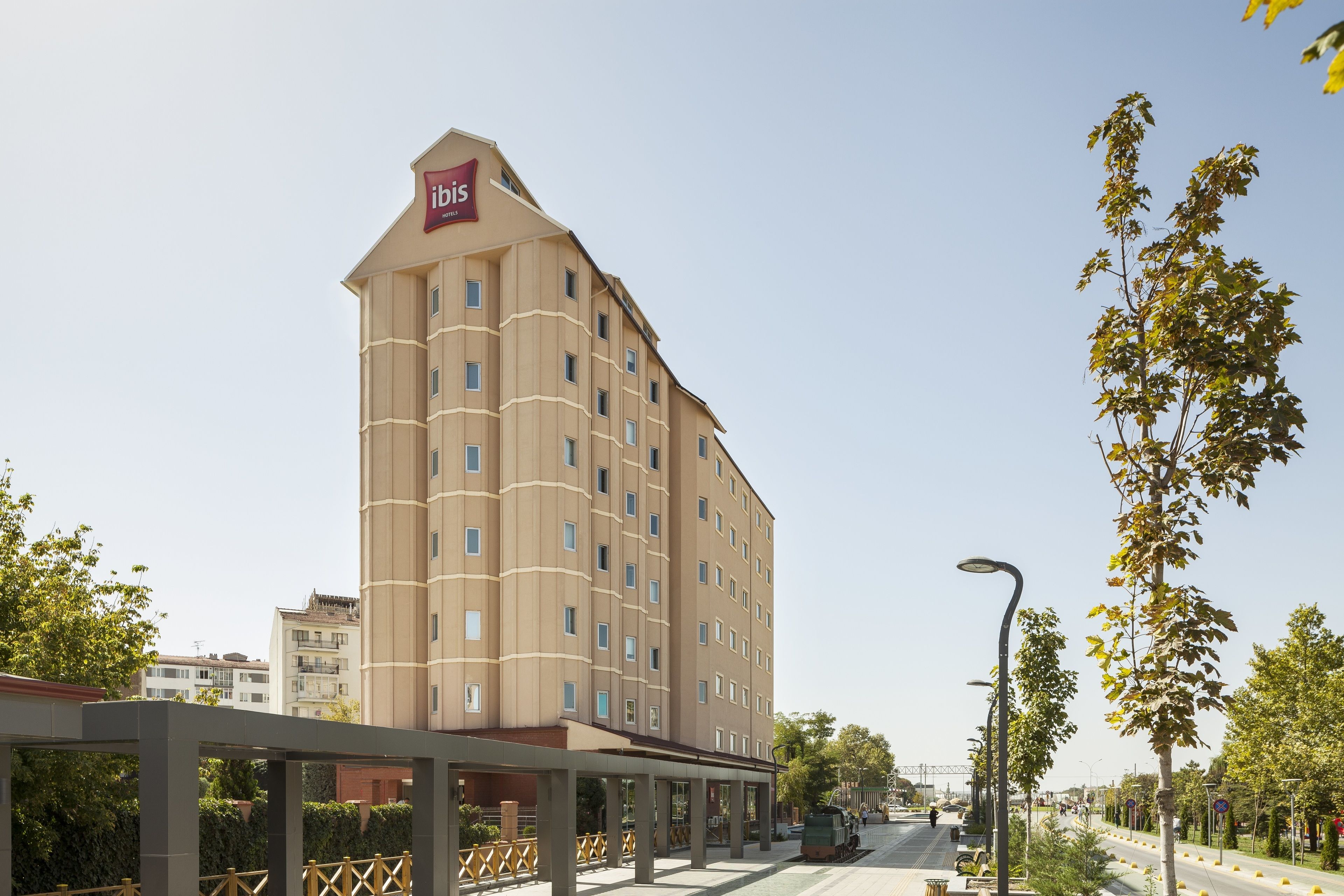 Un hotel de diverses plantes amb un senyal ben visible d'"ibis", una zona exterior coberta i un carrer amb arbres i topalls.