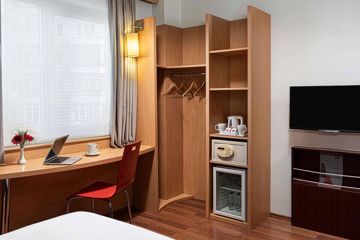 A hotel room with a wooden desk, a laptop, a red chair, a coat rack, a television, and a minibar.