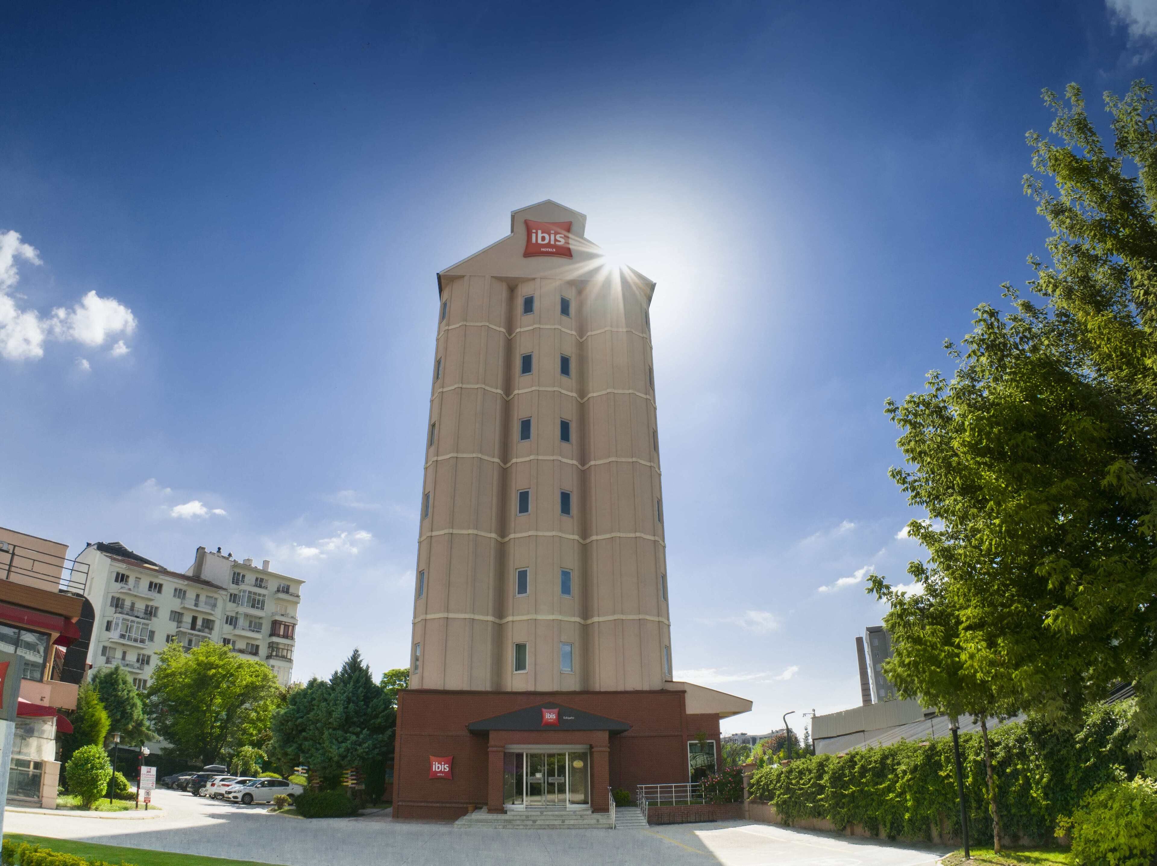 A tall, beige hotel with a prominent 'ibis' sign on the top, featuring a modern architectural design with a central core and multiple windows.