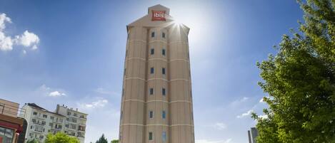 A tall, beige hotel with a prominent 'ibis' sign on the top, featuring a modern architectural design with a central core and multiple windows.
