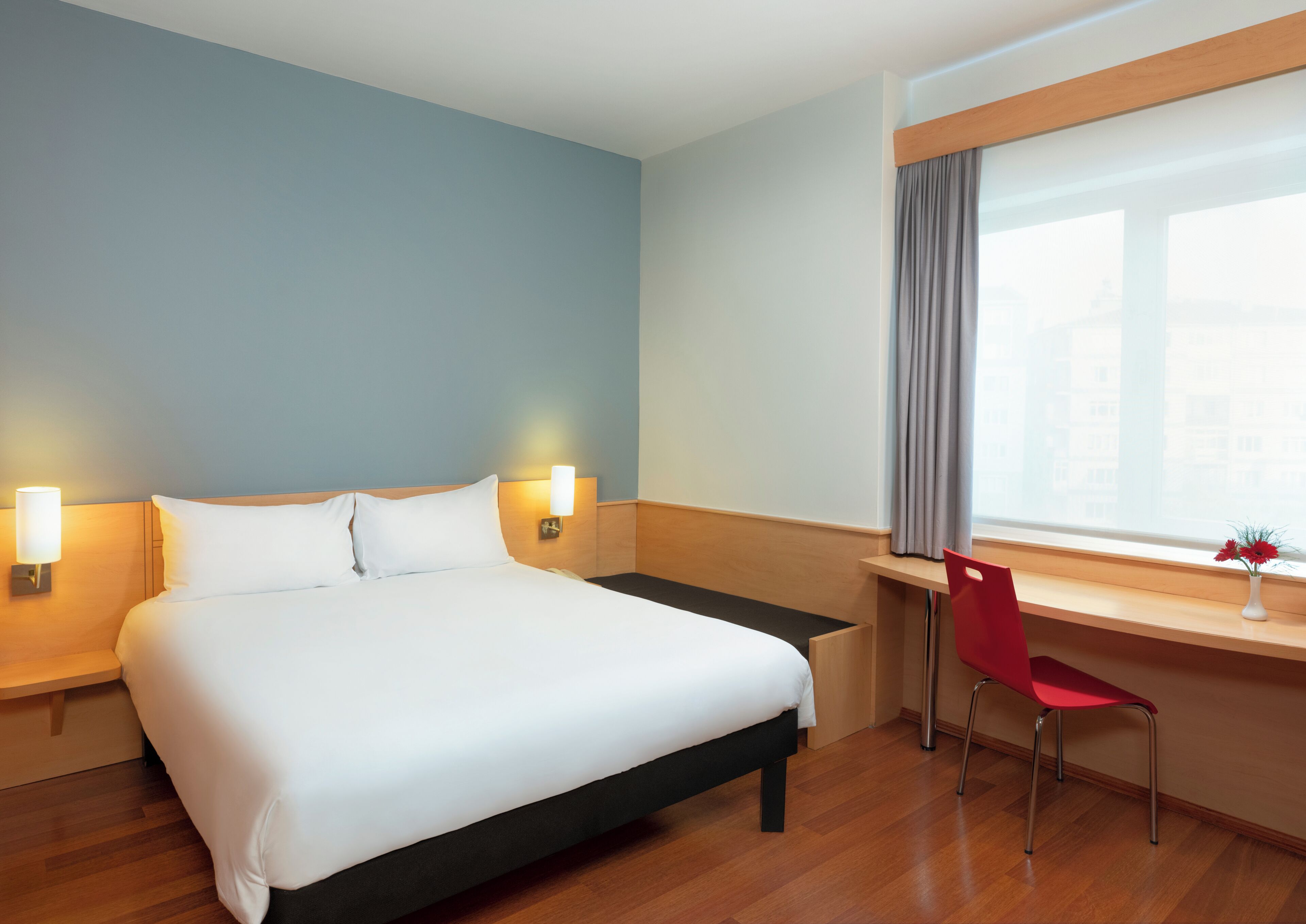 A hotel room with a bed, bedside lamps, a desk with a chair, a window with curtains, and a vase with flowers.