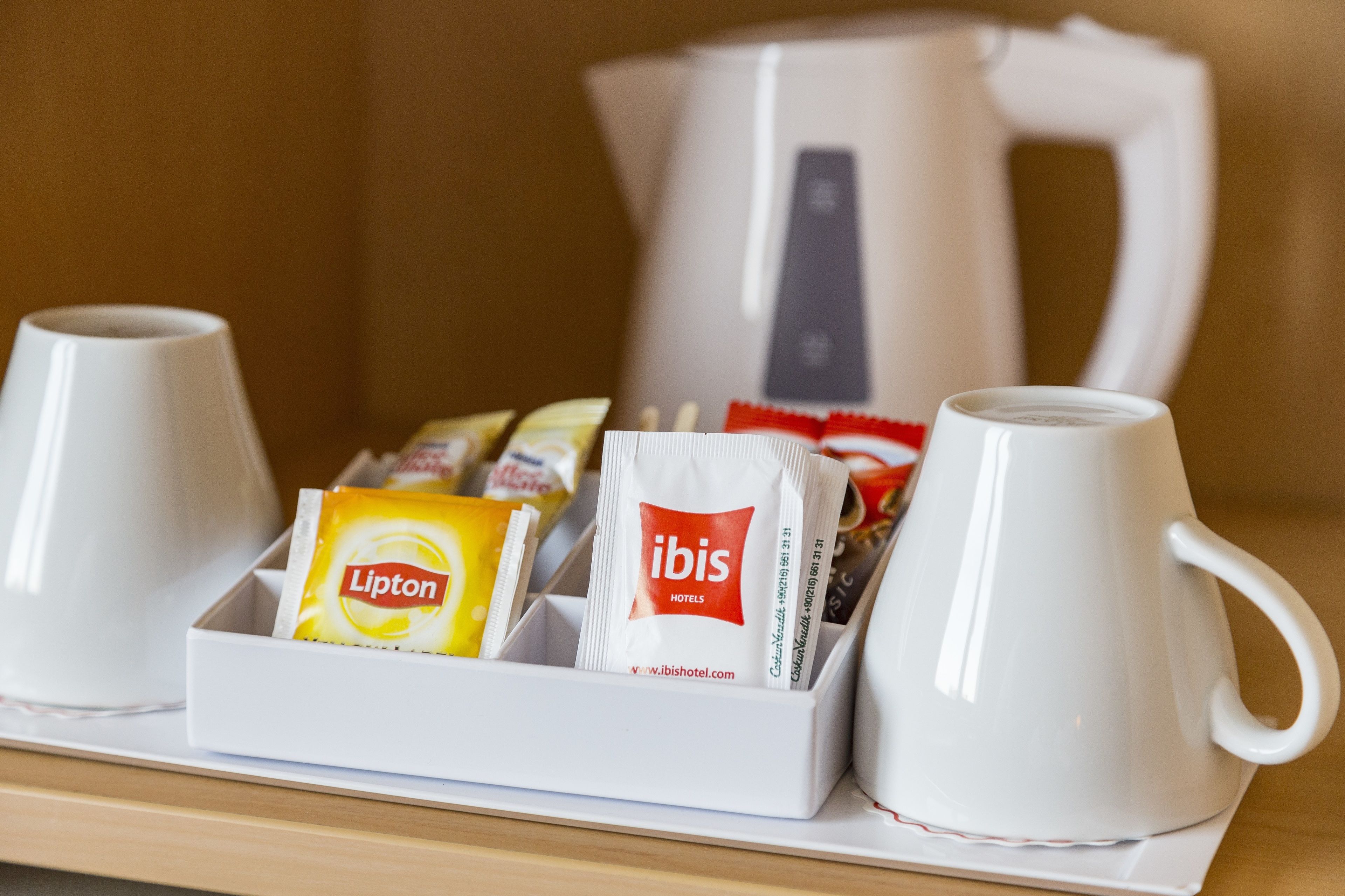Un plateau avec deux tasses blanches, une théière blanche et divers produits pour le petit-déjeuner, notamment du thé Lipton et des produits de la marque de l’hôtel Ibis.