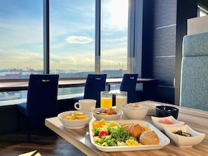 A breakfast table with a variety of dishes, including fruits, vegetables, and bread, set against a backdrop of a city view through large windows. - SOTETSU GRAND FRESA Shinagawa Seaside (Tokyo)