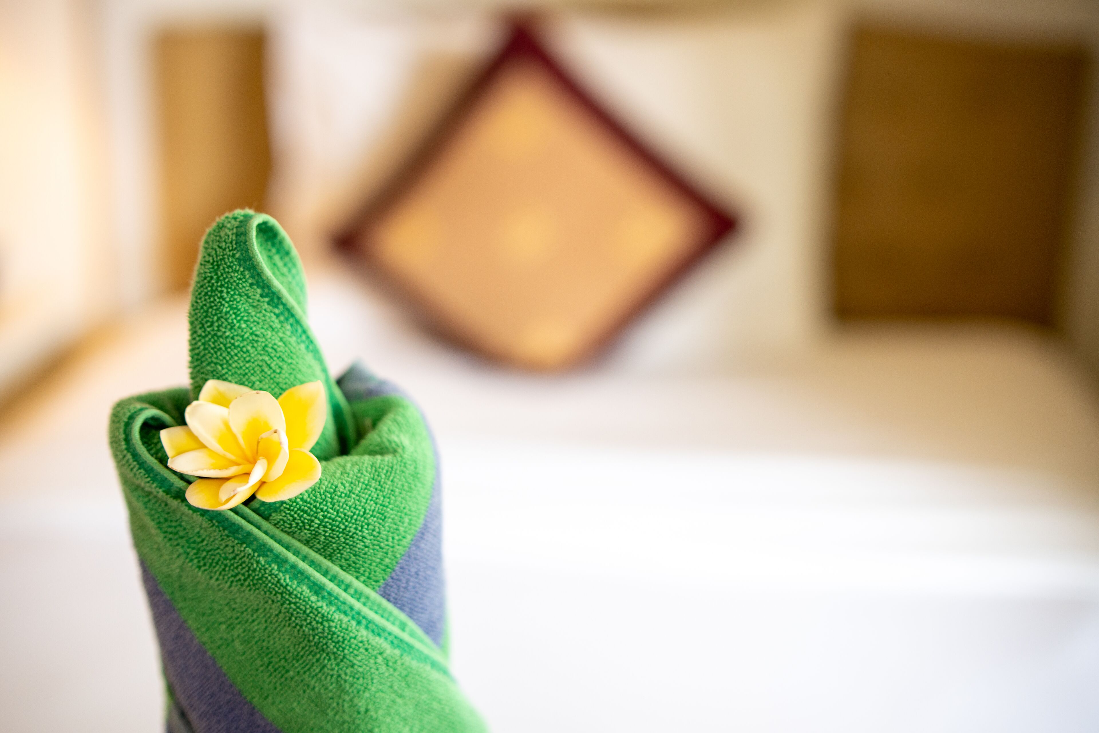 A neatly folded towel with a yellow flower, placed on a bed with a blurred background featuring a wooden headboard and a pillow.